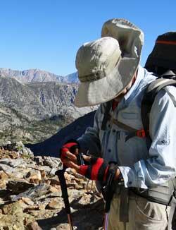 Katchan inserts one of his SEVEN batteries into his camera, Behind: steepest switchbacks of the trip on the east side of Bishop Pass